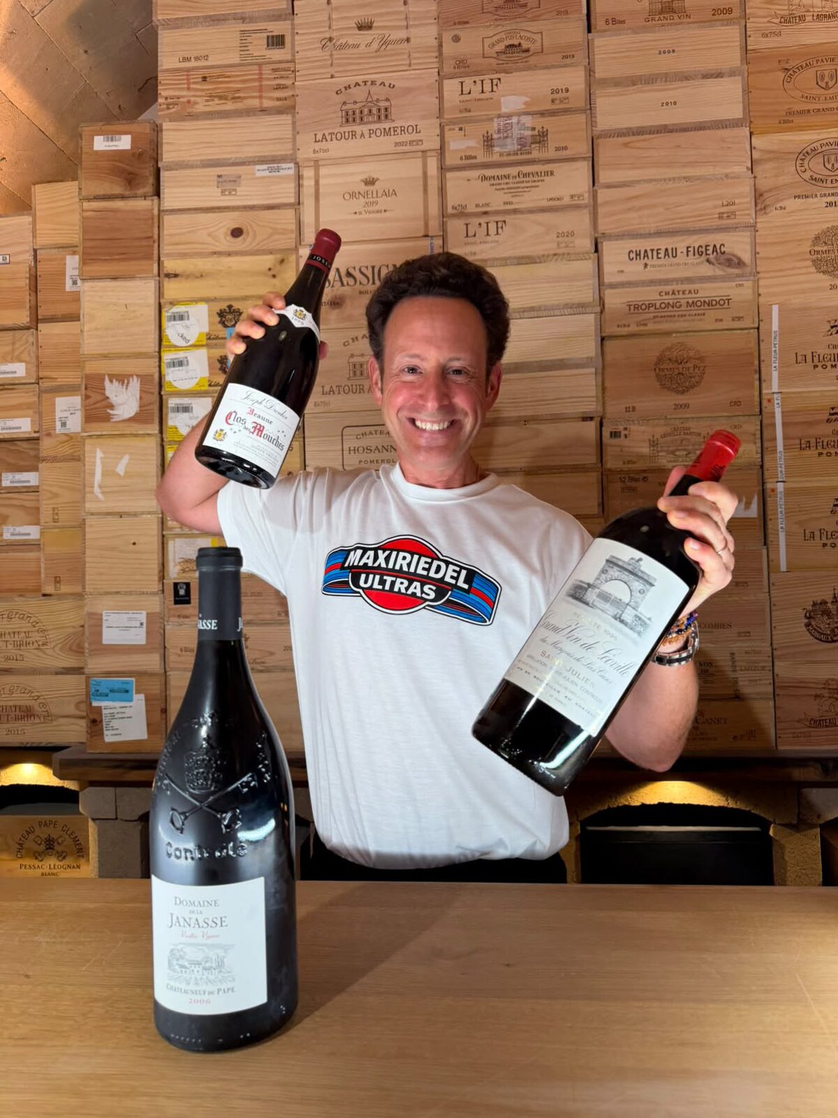Maximilian Riedel in his own wine cellar, wearing the white MaxiRiedelUltras T-shirt, in front of a wall of wooden wine cases. He raises a magnum of Joseph Drouhin Beaune Clos des Mouches in one hand and holds a magnum of Léoville Las Cases 1995 in the other; a magnum of Domaine de la Janasse Châteauneuf-du-Pape Vieilles Vignes 2006 stands on the counter in front of him.
