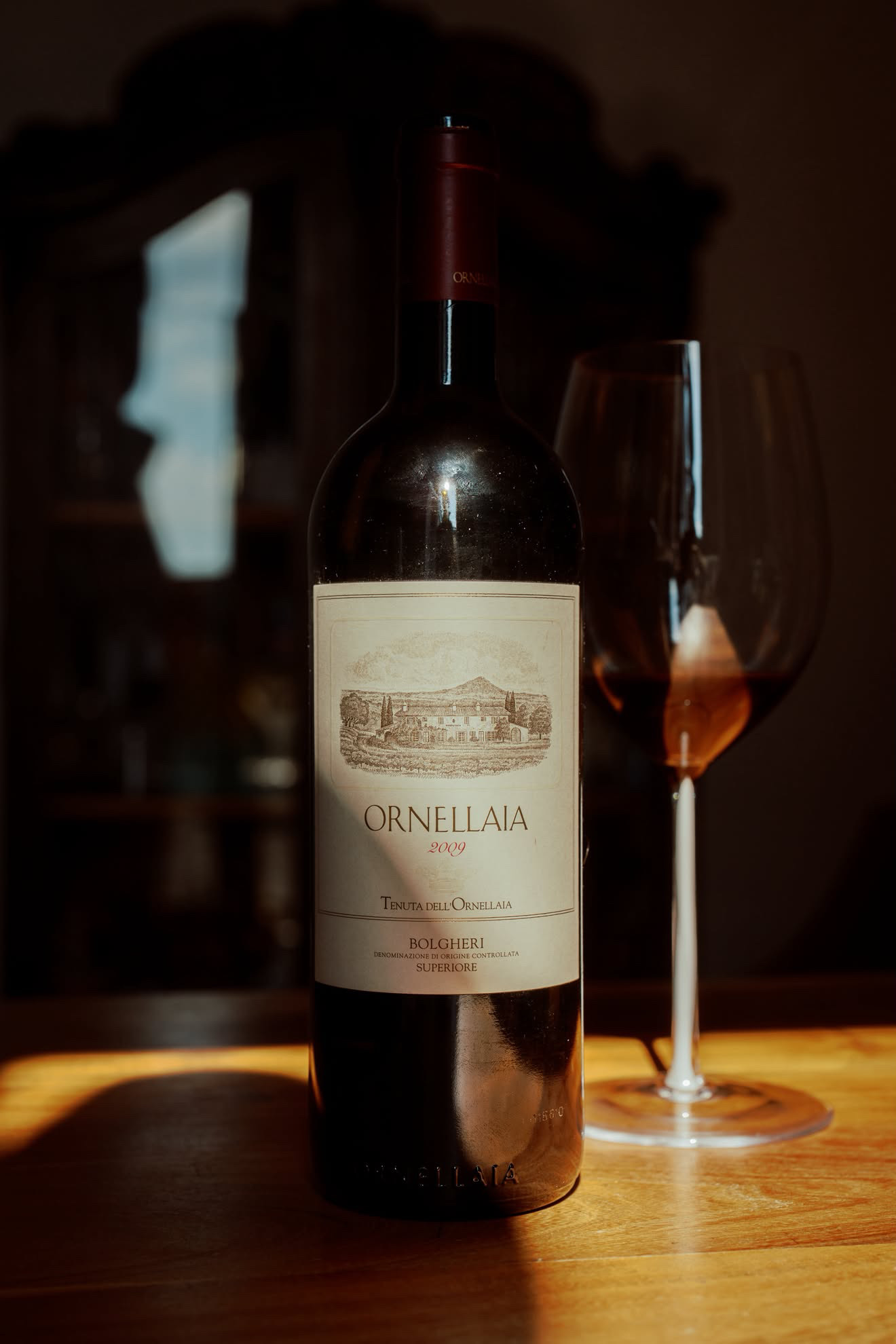 Ornellaia 2009 Bolgheri Superiore next to a Riedel Fatto a Mano Bordeaux glass with a white stem, a band of warm evening sun across the wooden table