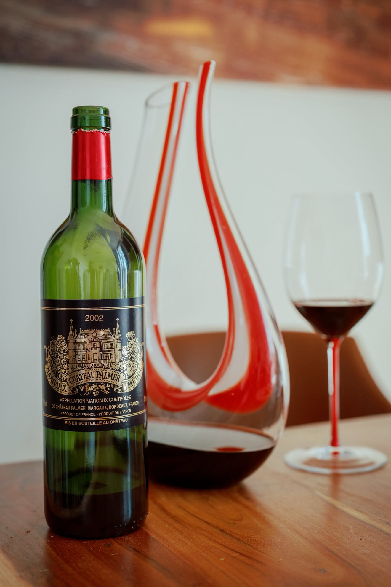 Ch&acirc;teau Palmer 2002 Margaux beside a Riedel Amadeo Fatto a Mano decanter with red ribbons and a Riedel Fatto a Mano Bordeaux glass with a red stem, on a wooden table